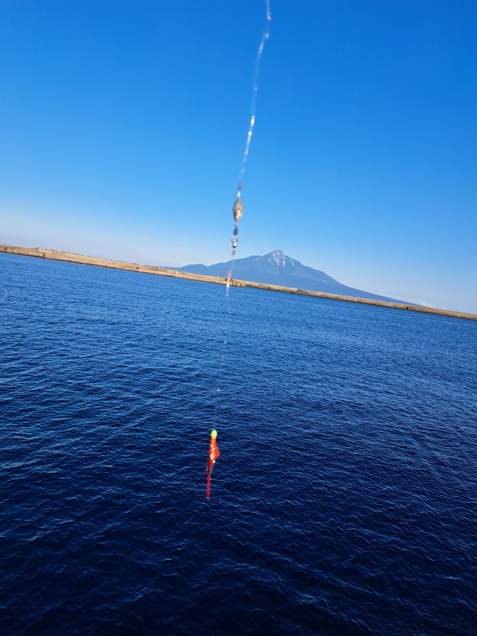 A fishing line and lure over deep blue water, with Mount Rishiri on the horizon